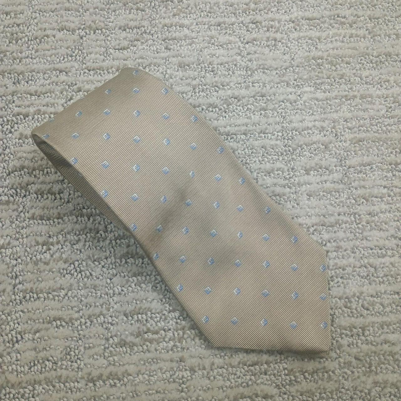 Brooks Brothers Makers necktie features a sophisticated tan or champagne ribbed silk base adorned with a neat geometric square pattern in light blue. This high-quality accessory was woven in England and expertly crafted in the USA from pure silk, ensuring a luxurious drape and classic professional look. The underside is finished with a signature sheep logo tipping, reflecting the brand's heritage of fine tailoring. Perfect for business meetings, weddings, or formal events, this tie offers a timeless aesthetic that pairs effortlessly with navy or charcoal suiting.