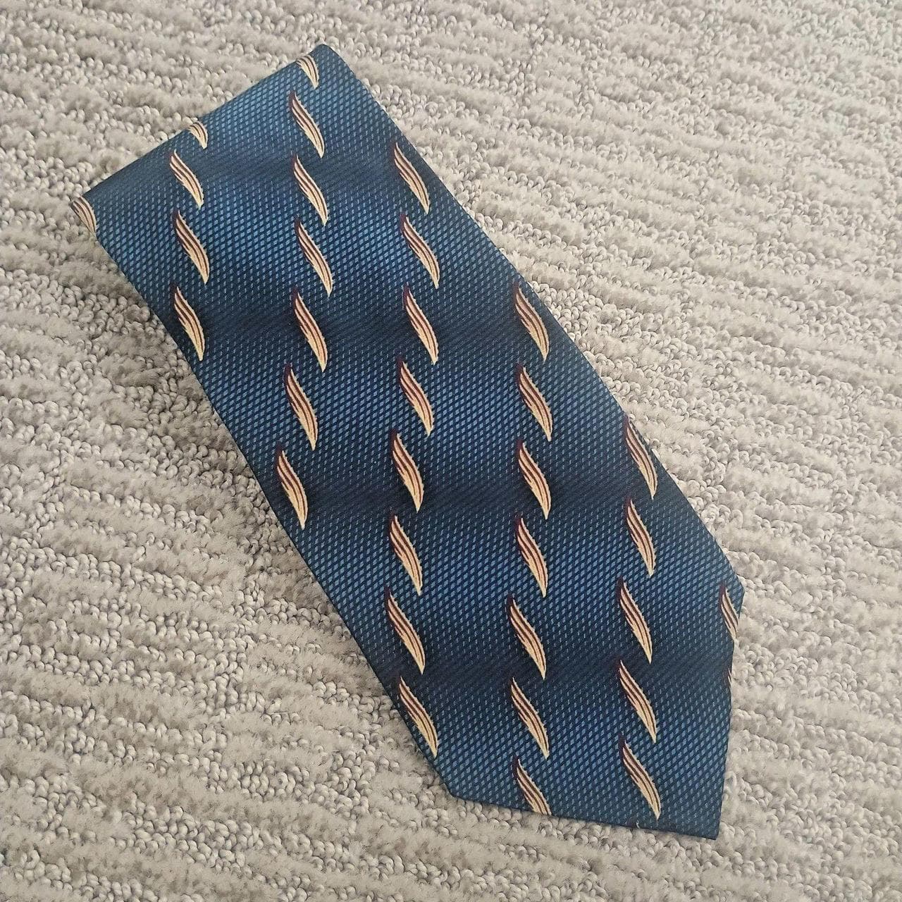 Bill Blass Black Label Silk Necktie, Blue with Tan Leaf Pattern. This elegant necktie features a textured blue base accented by a repeat leaf motif in tan and burgundy tones.